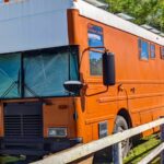 Bright orange converted bus with white roof parked outdoors next to wooden fence, surrounded by trees, with reflective covering on front windshield. Post is about the converted bus interior tour.