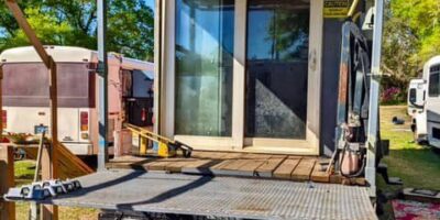 Open back deck attached to converted bus with metal railing, small table, storage bins, and sliding glass doors leading into the interior.