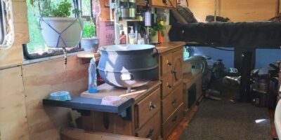 Converted bus kitchen with wooden cabinetry, sink under curtained window, produce basket, countertop appliances, and a view into built-in shelving and sleeping loft above driver’s area.
