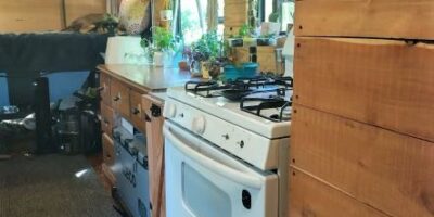 Converted bus kitchen with full-size white stove, wood countertops, overhead cabinets with baskets, hanging plants by curtained windows, and wood-paneled ceiling.