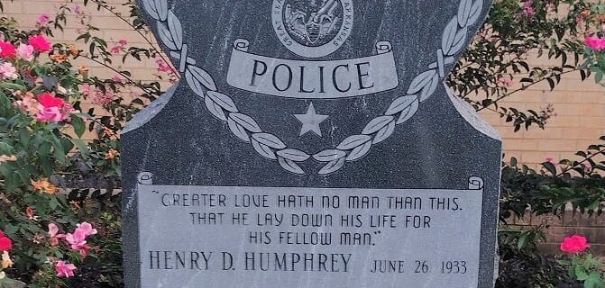 Henry D. Humphrey memorial, Stone, shaped with a heart-like top and engraved eagle. Center badge emblem encircled by laurel wreath. Inscription reads: “Greater love hath no man than this, that he lay down his life for his fellow man.” Surrounded by blooming rose bushes, placed in front of a brick wall. Honors a fallen Alma, Arkansas police officer killed in the line of duty.