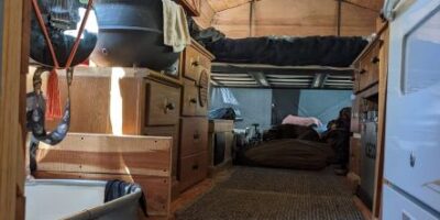 Converted bus kitchen with wood-paneled ceiling, carpeted floor, hanging black pot, wooden cabinetry on both sides, and compact white appliance beside drawers