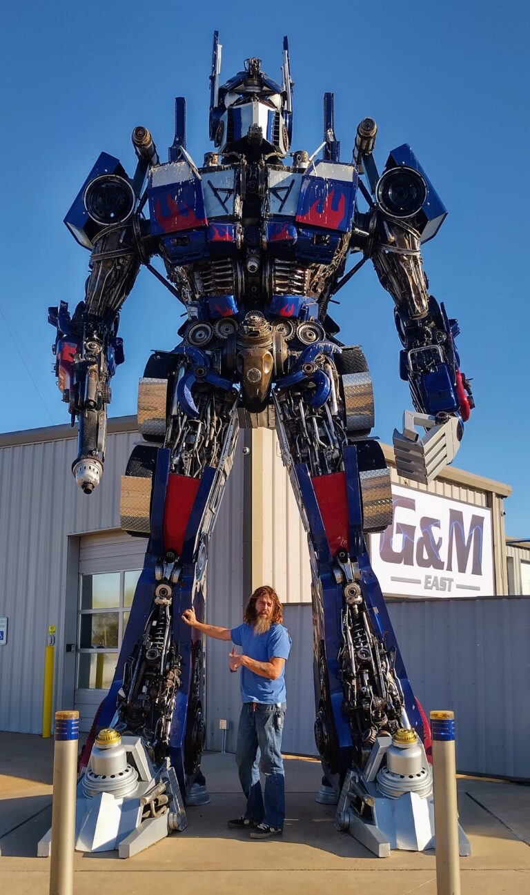 Optimus Prime statue at G&M Body Shop in Stillwater, Oklahoma, with a person posing in front, arm extended toward the towering robot. Constructed from metal parts and labeled “G&M GIANT” in the background, this figure anchors the east side of the Transformers statues Stillwater Oklahoma display.