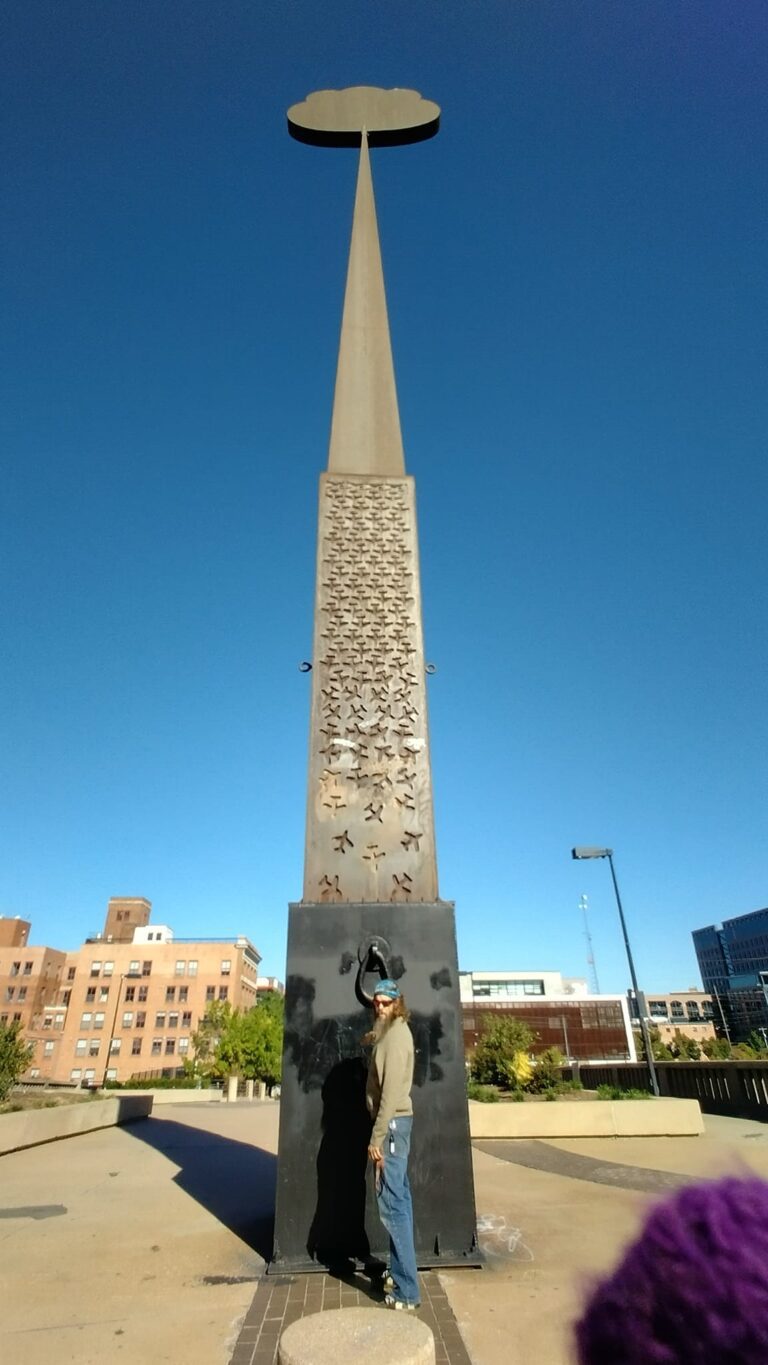 Triangular monument in downtown Tulsa with a flat disc top, inscribed vertically in multiple languages, with a person standing at its base; trees, buildings, and a clear blue sky in the background — part of the Center of the Universe Tulsa area.