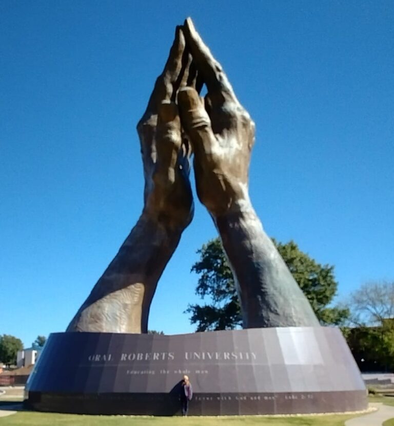 Wide shot of the world's largest praying hands sculpture at Oral Roberts University, with bronze hands clasped in prayer atop a grassy lawn. A person stands at the base for scale. Trees and a clear blue sky frame the outdoor setting. “ORAL ROBERTS UNIVERSITY” is inscribed on the pedestal.