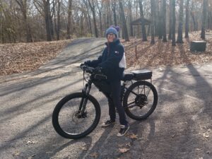 Bus Life Break Maker: Nancy standing beside a black e-bike on a paved path, surrounded by autumn leaves and trees, wearing a blue beanie and dark jacket.