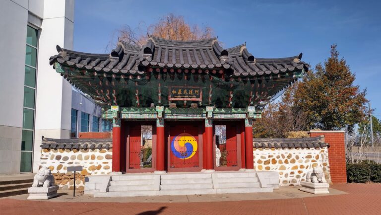 Traditional Korean-style gate at the H.U. Lee International Gate and Garden in Little Rock, Arkansas, featuring a curved tiled roof, vibrant red doors with a Sam-Taegeuk symbol, flanked by stone lion statues and patterned stone walls, with autumn trees and city buildings in the background.