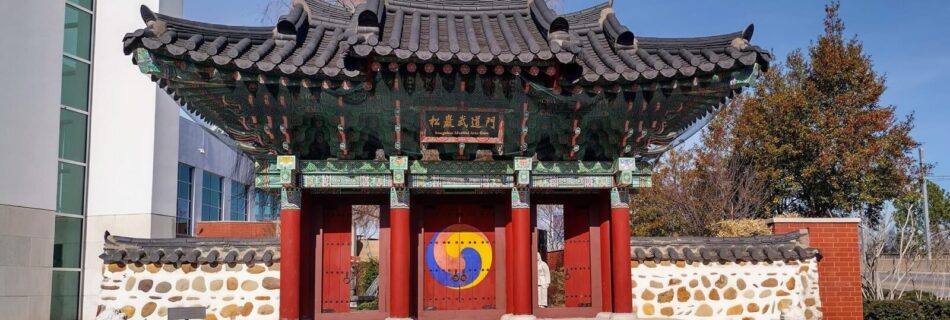 Traditional Korean-style gate at the H.U. Lee International Gate and Garden in Little Rock, Arkansas, featuring a curved tiled roof, vibrant red doors with a Sam-Taegeuk symbol, flanked by stone lion statues and patterned stone walls, with autumn trees and city buildings in the background.