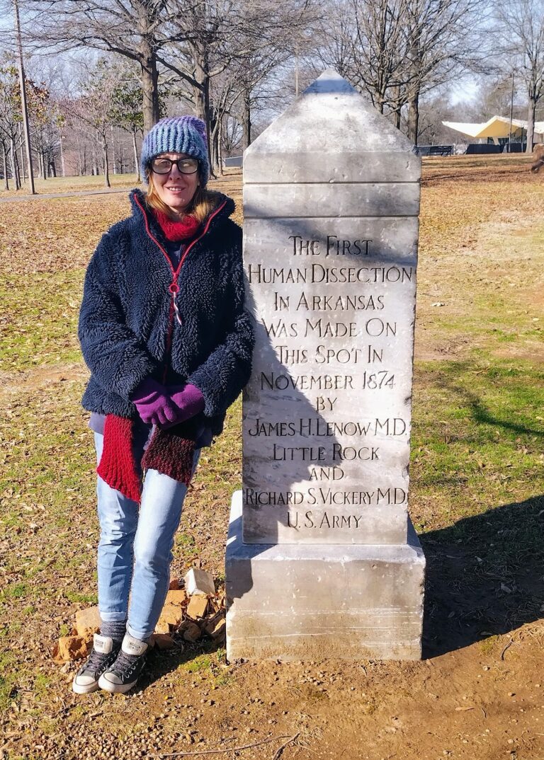 Person in winter gear standing beside the Human Dissection Monument in MacArthur Park — granite obelisk with engraved inscription marking Arkansas’s first legal dissection in 1872.