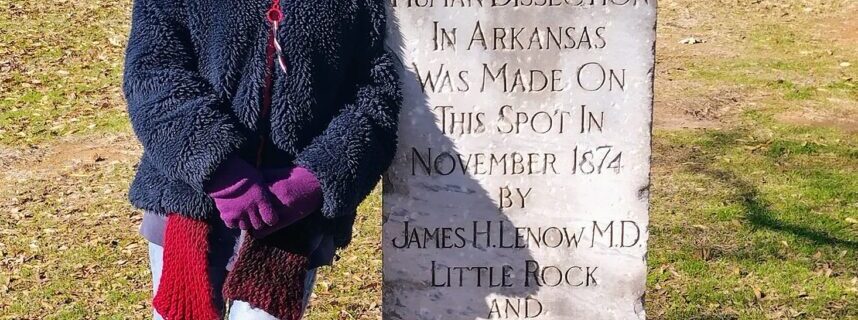 Person in winter gear standing beside the Human Dissection Monument in MacArthur Park — granite obelisk with engraved inscription marking Arkansas’s first legal dissection in 1872.
