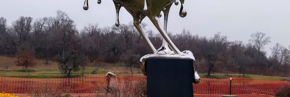 Mirrored bird sculpture titled Reflection in Nashville’s Shelby Park—appears to melt from the pedestal with metallic drips, set in a grassy area with red fencing and trees in the background