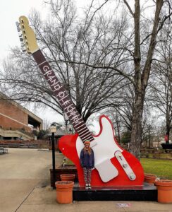 Person standing in front of the red and white Grand Ole Opry guitar statue outdoors, with leafless trees and a brick building in the background.
