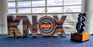 Wooden sculpture spelling “KNOX” with a circular Knoxville sign inside the “O,” and a statue of a figure on a pedestal beside it, framed by convention center windows