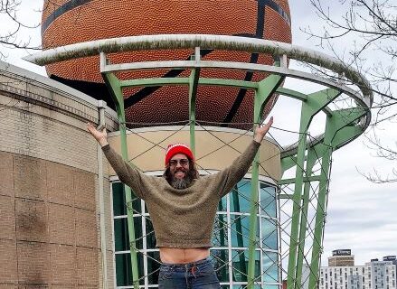 A person stands in front of the World's Largest Basketball sculpture in Knoxville, Tennessee, raising their arms to mimic the hoop structure above. The oversized fiberglass basketball is integrated into the building's architecture, appearing as if it is going through a metal hoop. Trees and a cityscape with buildings are visible in the background.