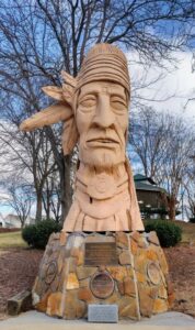 Peter Wolf Toth's 'Junaluska' Whispering Giant sculpture in Johnson City, Tennessee - a carved wooden Native American figure with traditional headband and detailed facial features, mounted on a stone base with commemorative plaques featured on a page about the Trail of the Whispering Giants