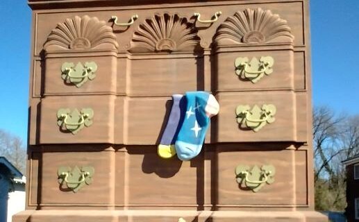 The World's Largest Chest of Drawers, a towering 38-foot furniture landmark in High Point, North Carolina. Its ornate gold handles and playful oversized socks hanging from an open drawer highlight the city's rich furniture heritage. The bold lettering reads 'Furniture Capital of the World,' reinforcing its iconic status. A visitor stands in front, showing its impressive scale.