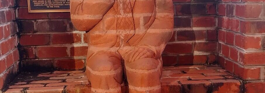 A life-sized brick sculpture of a seated man integrated into the wall, dressed in a suit and tie, with his right hand resting as if holding a small object. A commemorative plaque mounted above the sculpture provides historical context, though the text is not fully visible in this image.