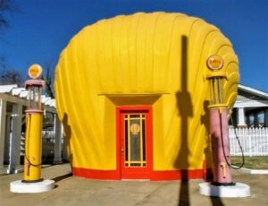 The Last Shell-Shaped Shell Service Station, a bright yellow, seashell-shaped gas station with red accents, featuring two vintage Shell-branded gas pumps—one yellow and one pink—standing in front, enclosed by a white fence under a clear blue sky.