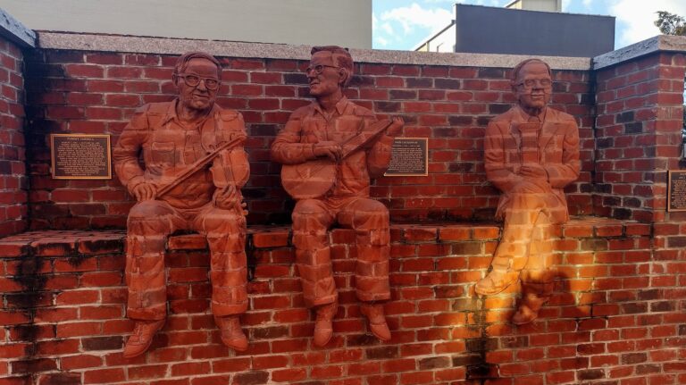Three life-sized brick sculptures of musicians seamlessly integrated into a brick wall, each holding a different instrument—two with guitars and one with a banjo. The textured brick material makes the figures appear as if emerging from the wall itself, creating a striking, immersive effect. Two plaques mounted between the sculptures provide historical or artistic context for the installation.