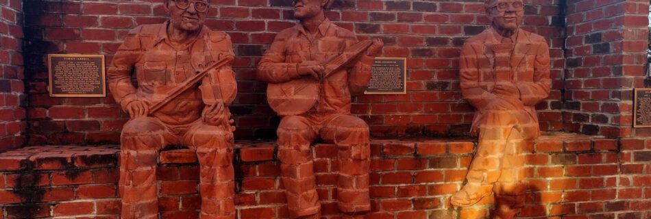 Three life-sized brick sculptures of musicians seamlessly integrated into a brick wall, each holding a different instrument—two with guitars and one with a banjo. The textured brick material makes the figures appear as if emerging from the wall itself, creating a striking, immersive effect. Two plaques mounted between the sculptures provide historical or artistic context for the installation.