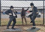 Statues of Jake and Elwood Blues from The Blues Brothers, dressed in black suits, sunglasses, and black hats, standing in front of a chain-link fence. Jake is posed adjusting his tie, while Elwood strikes a mid-dance stance. Behind them, Zach stands playfully in the background, partially visible through the fence, adding an extra layer of personality to the scene. Captured outdoors in Stedman, NC.
