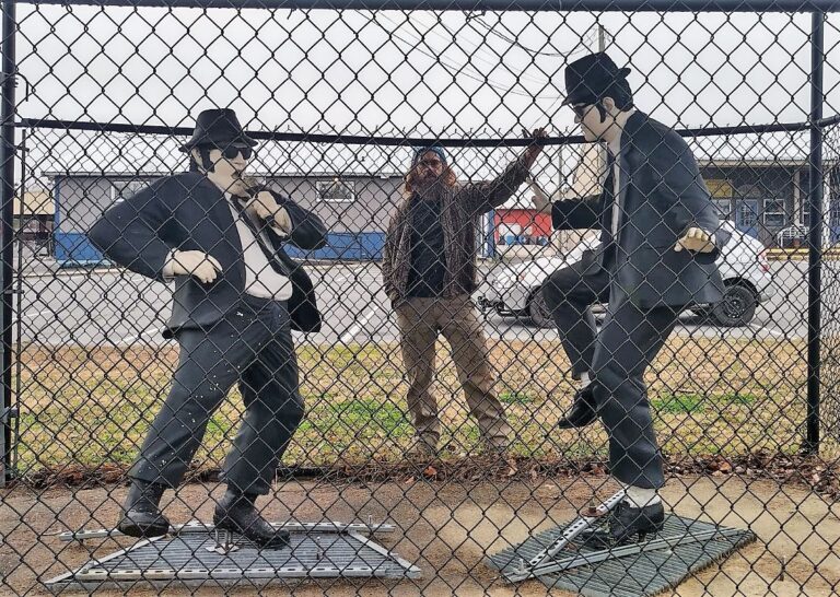 Statues of Jake and Elwood Blues from The Blues Brothers, dressed in black suits, sunglasses, and black hats, standing in front of a chain-link fence. Jake is posed adjusting his tie, while Elwood strikes a mid-dance stance. Behind them, Zach stands playfully in the background, partially visible through the fence, adding an extra layer of personality to the scene. Captured outdoors in Stedman, NC.
