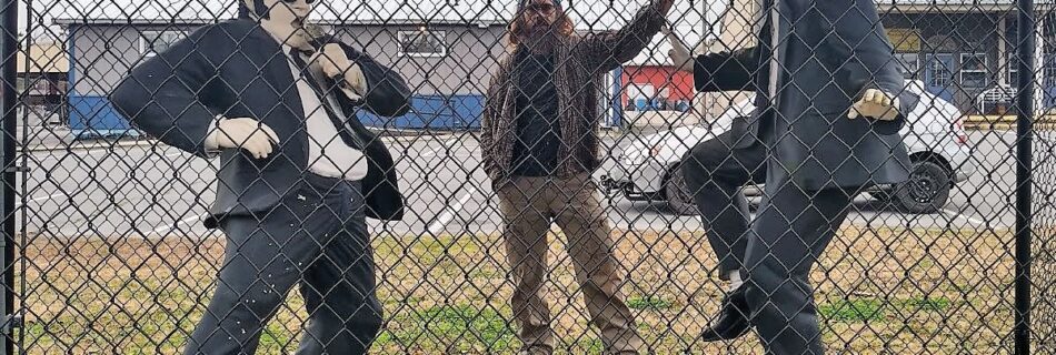 Statues of Jake and Elwood Blues from The Blues Brothers, dressed in black suits, sunglasses, and black hats, standing in front of a chain-link fence. Jake is posed adjusting his tie, while Elwood strikes a mid-dance stance. Behind them, Zach stands playfully in the background, partially visible through the fence, adding an extra layer of personality to the scene. Captured outdoors in Stedman, NC.