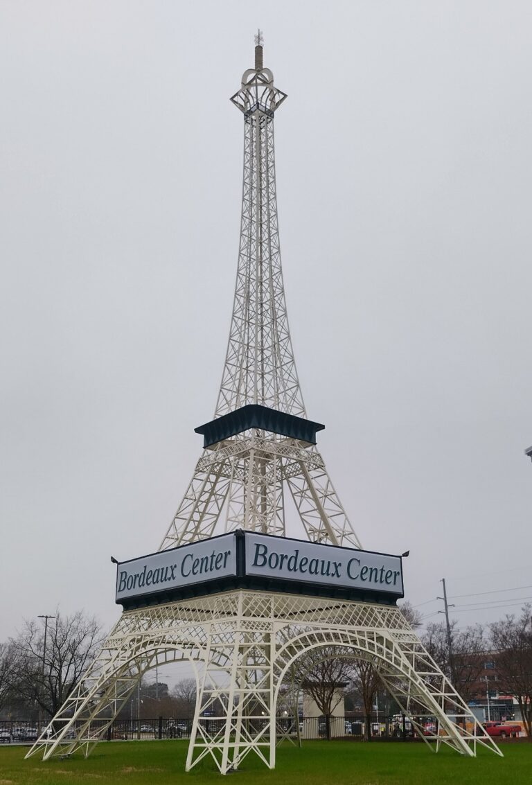Miniature white Eiffel Tower replica standing in a commercial parking lot at Bordeaux Center in Fayetteville, North Carolina, framed by a blue sky and shopping plaza backdrop.