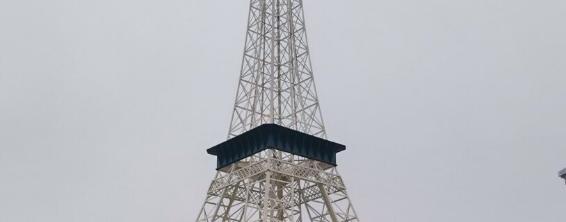 Miniature white Eiffel Tower replica standing in a commercial parking lot at Bordeaux Center in Fayetteville, North Carolina, framed by a blue sky and shopping plaza backdrop.