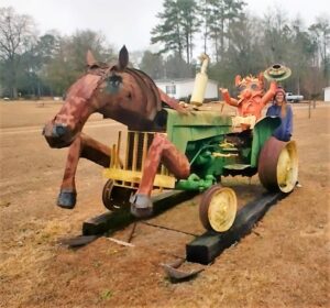 Sculpture of a hybrid creature with the front half resembling a painted horse and the rear half built from a green tractor, with yellow wheels and a seated mannequin posed as the driver. A person stands beside it for scale, with trees and buildings in the background.
