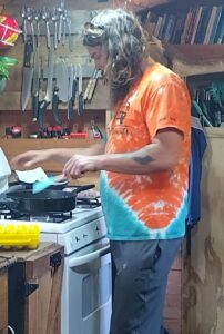 Zach cooking in the bus kitchen, wearing a tie-dye shirt with his long hair down, focused on a stovetop pan while surrounded by hanging utensils and a shelf of books—bus life at its coziest.