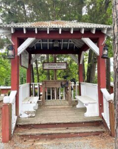 Wooden gazebo with white benches and red trim at the Angus Barn, flanked by lanterns and trees, offering a quiet resting spot beside the chaos of the Kicking Booth.