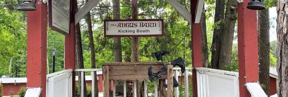Wooden gazebo with white benches and red trim at the Angus Barn, flanked by lanterns and trees, offering a quiet resting spot beside the chaos of the Kicking Booth.
