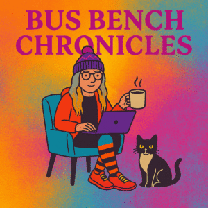 Nancy writing from her Bus Bench Chronicles setup: stocking cap on, El Macké open, coffee close by, and her black cow kitty curled at her feet—snapshot from the Michigan Bus Break