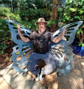 Zach sitting on a metallic butterfly wing bench in a garden, wearing a black graphic tee and white sneakers, blending nature with roadside whimsy. At Reiman Gardens Ames Iowa