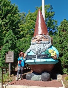 A visitor poses beside a towering concrete gnome holding a yellow flower at Reiman Gardens in Ames, Iowa, surrounded by lush greenery. A nearby welcome sign highlights Reiman Gardens as one of the whimsical things to do in Ames, Iowa