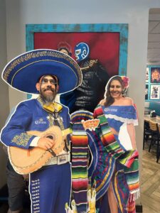 Nancy and Zach posing with life-sized cardboard mariachi and folklórico cutouts inside a Mexican restaurant—Corona logos, luchador masks, and Nancy holding the chaos together while Zach mugs for the camera.