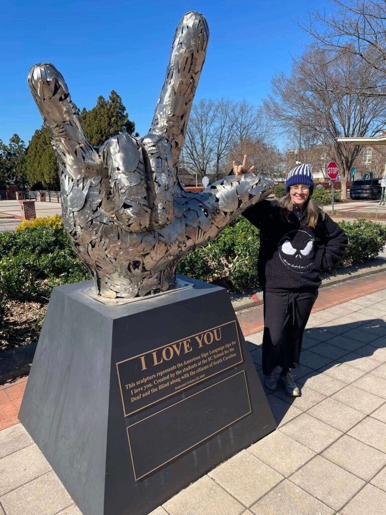 Towering metallic sculpture of a hand making the “I love you” gesture in American Sign Language. Mounted on a pedestal with a plaque reading “I LOVE YOU,” the piece celebrates inclusivity and connection. A person in a black outfit and beanie mirrors the sign beside it, adding a human echo. Set against a backdrop of trees and a bright blue sky along a paved walkway, the scene feels heartfelt and iconic