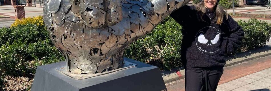 Towering metallic sculpture of a hand making the “I love you” gesture in American Sign Language. Mounted on a pedestal with a plaque reading “I LOVE YOU,” the piece celebrates inclusivity and connection. A person in a black outfit and beanie mirrors the sign beside it, adding a human echo. Set against a backdrop of trees and a bright blue sky along a paved walkway, the scene feels heartfelt and iconic