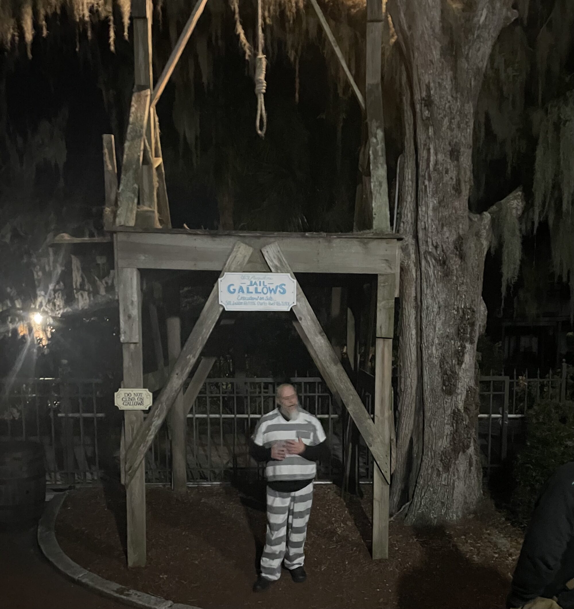 Tour guide in striped prison costume standing under a wooden gallows at night, with a noose overhead and trees in the background.