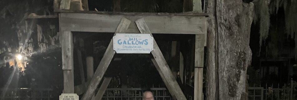 Tour guide in striped prison costume standing under a wooden gallows at night, with a noose overhead and trees in the background.