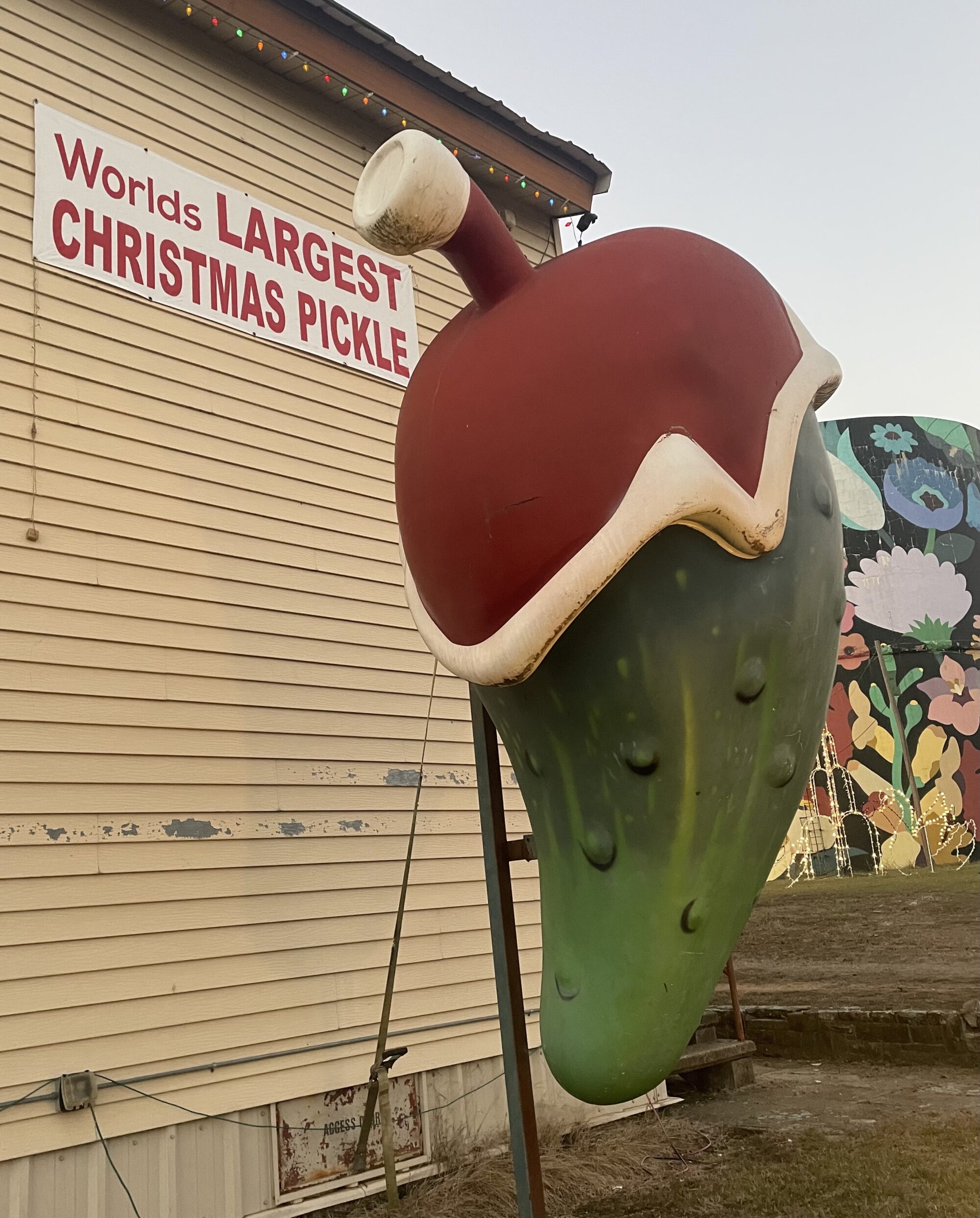 The World’s Largest Christmas Pickle statue wearing a Santa hat at Christmas Winter Wonderland in Fort Smith, Arkansas, photographed during dusk with the building and sign in the background.