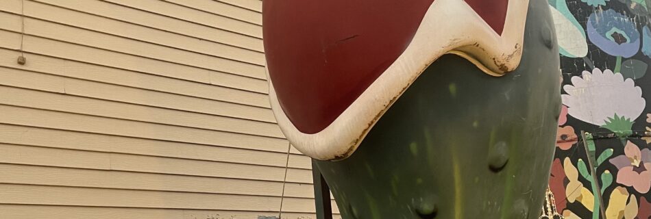 The World’s Largest Christmas Pickle statue wearing a Santa hat at Christmas Winter Wonderland in Fort Smith, Arkansas, photographed during dusk with the building and sign in the background.