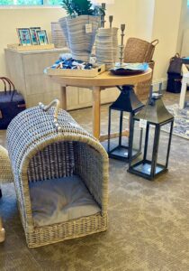 A wicker pet house with an arched entrance and cushioned interior, displayed at Furnitureland South next to a round table set with home decor and lanterns — the one we nicknamed 'Phil's Future Headquarters'