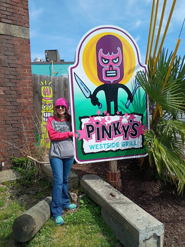 A woman (Nancy) stands beside the outdoor sign for Pinky's Westside Grill in Charlotte, NC, smiling at the camera. The illustrated sign features a cartoon luchador wrestler character holding a knife against a yellow and green circular background with the restaurant name in bold lettering. She is wearing a pink baseball cap, sunglasses, a Star Wars hoodie, and jeans. A brick wall, colorful murals, and palm-like plants are visible in the background.