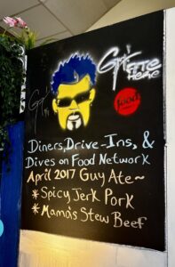 Chalkboard inside Carib Shack showing Guy Fieri's portrait and the Diners, Drive-Ins and Dives dishes he ordered during his 2017 visit.