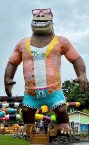 The full-body view of the giant 'Hugh Mongous' statue towering over a water park entrance, with visitors standing at its feet for scale.