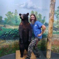 Zach “hugging the wildlife” with a life‑sized taxidermy bear at Call of the Wild Museum in Gaylord, Michigan, against a painted forest backdrop.