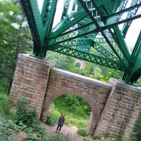 Person standing beneath a large green steel bridge supported by stone pillars, framed through an arched base, with lush forest greenery surrounding the structure.