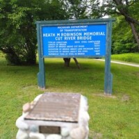 Informational sign for the Heath M. Robinson Memorial Cut River Bridge in Michigan, with a stone monument in front and trees in the background.